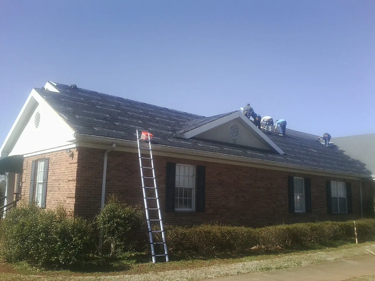 Crew tearing shingles off a brick ranch roof with a ladder set — Emergency Roof Repair work by our crew in Tama