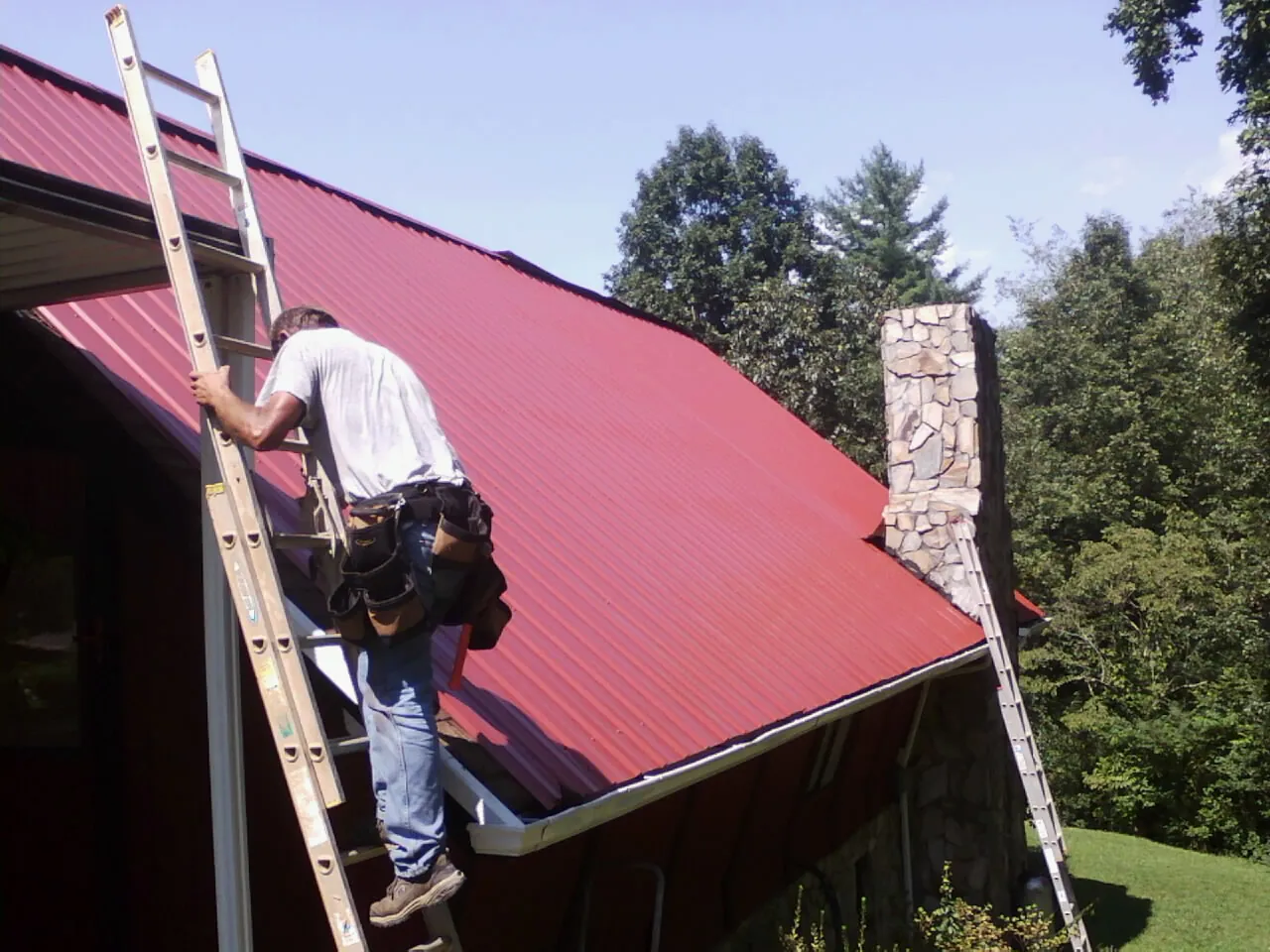 Roofer on a ladder beside a red metal roof and stone chimney — Soffit & Fascia Repair by Roofing Brothers in Tama